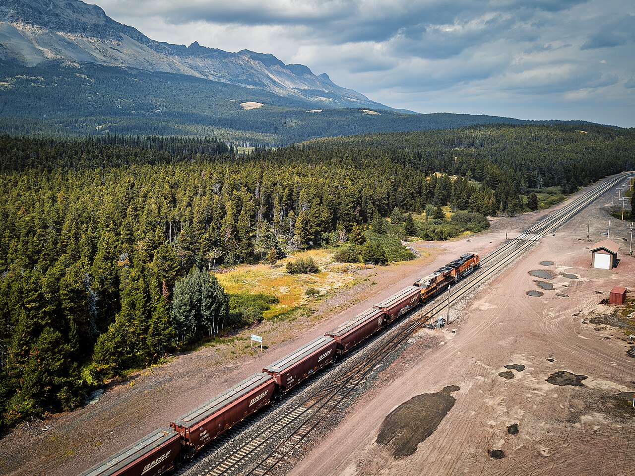 An aerial view of Marias Pass on the Continental Divide, where a freight train threads through the forested Montana Rockies alongside US-2.